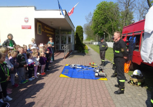Dzieci stoją na dworze wokół wozu strażackiego, strażacy prezentują narzędzia znajdujące się w wozie strażackim.